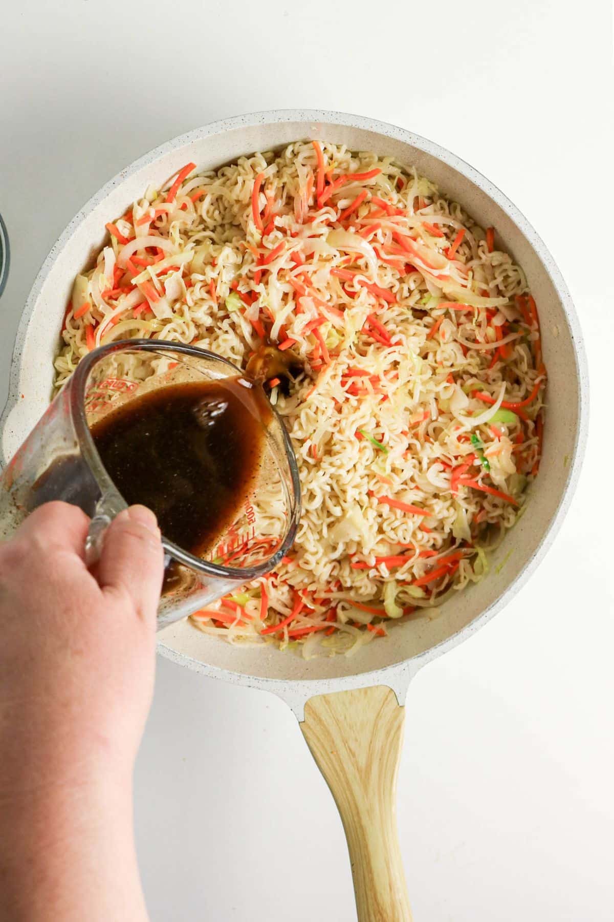 Hand pouring dark sauce over noodles, shredded carrots, and cabbage in a white pan on a white background.