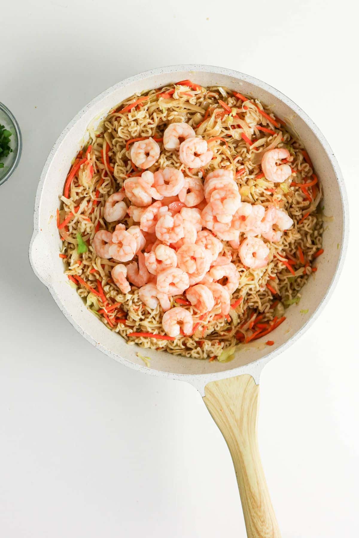 A skillet of cooked ramen noodles with vegetables topped with shrimp on a white surface.