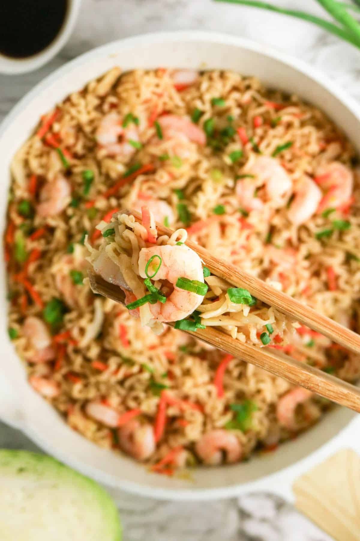 Chopsticks holding shrimp and noodles over a bowl of shrimp stir-fry with vegetables, garnished with green onions.