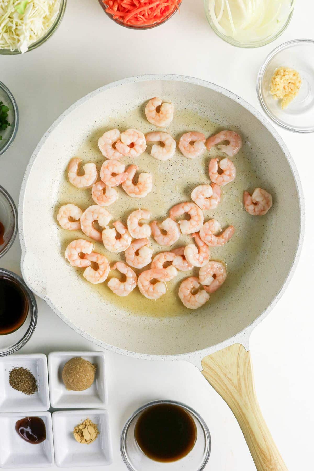 Raw shrimp cooking in a white skillet, surrounded by bowls of sauces, spices, and chopped vegetables.