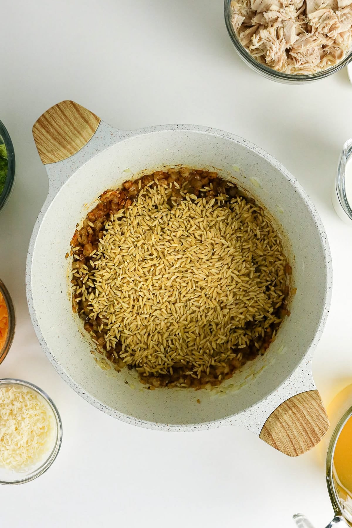 White pot with uncooked brown rice and seasonings, surrounded by bowls of shredded chicken, cheese, and other ingredients.