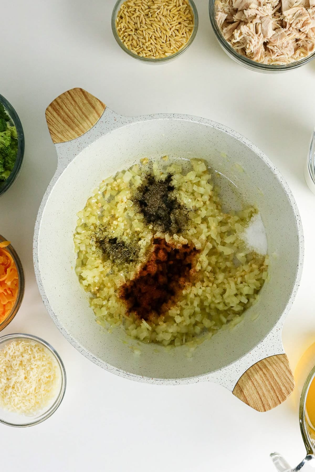 Diced onions and spices cooking in a white pot, surrounded by bowls of ingredients on a white surface.