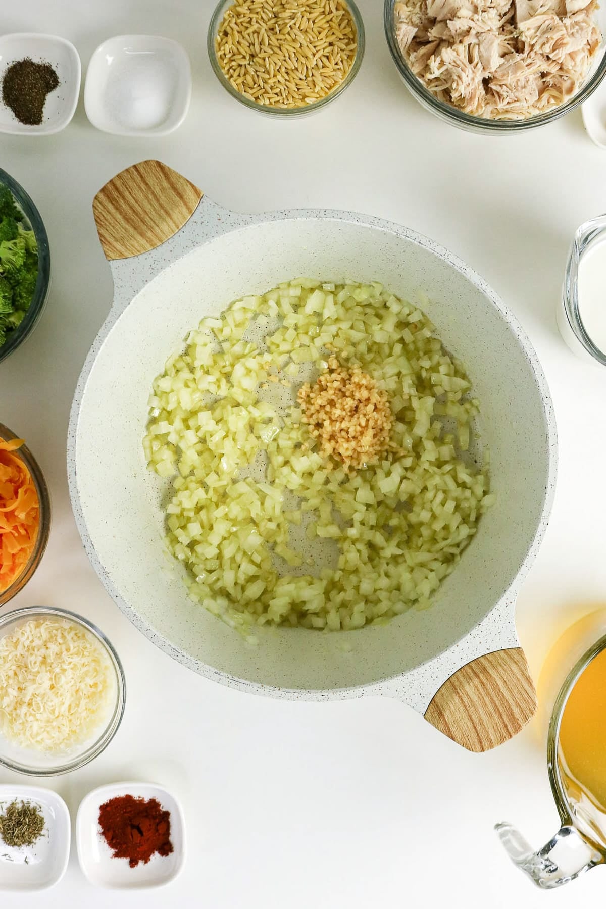 Overhead view of chopped onions and minced garlic sautéing in a white pot, surrounded by soup ingredients.