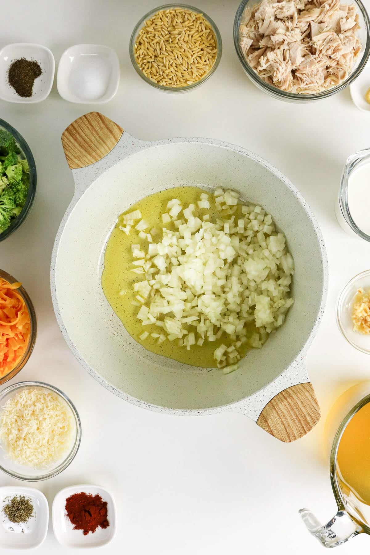 Chopped onions cooking in a white pot, surrounded by bowls of pasta, shredded chicken, cheese, veggies, and spices.