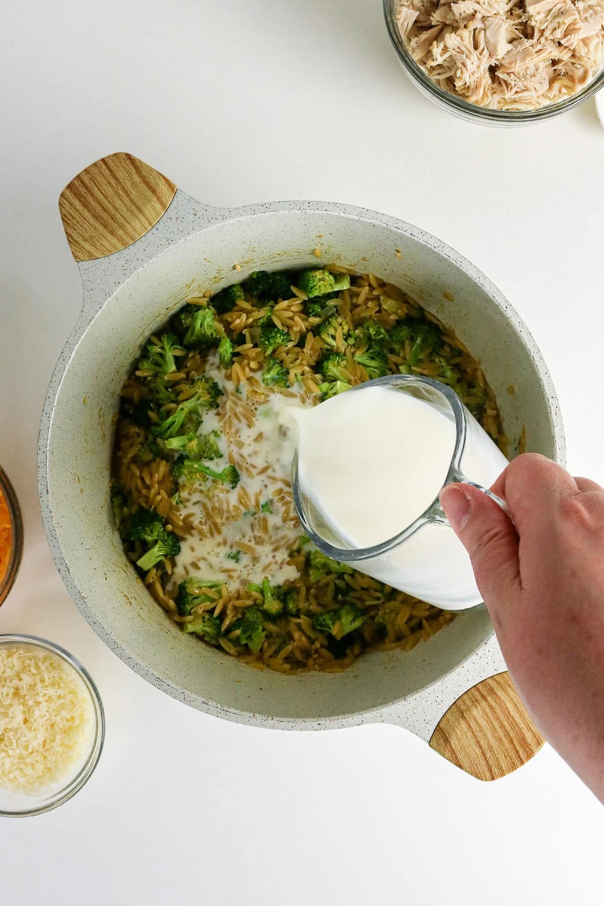 A hand pours milk into a pot of orzo and broccoli, surrounded by bowls of cheese and shredded chicken.