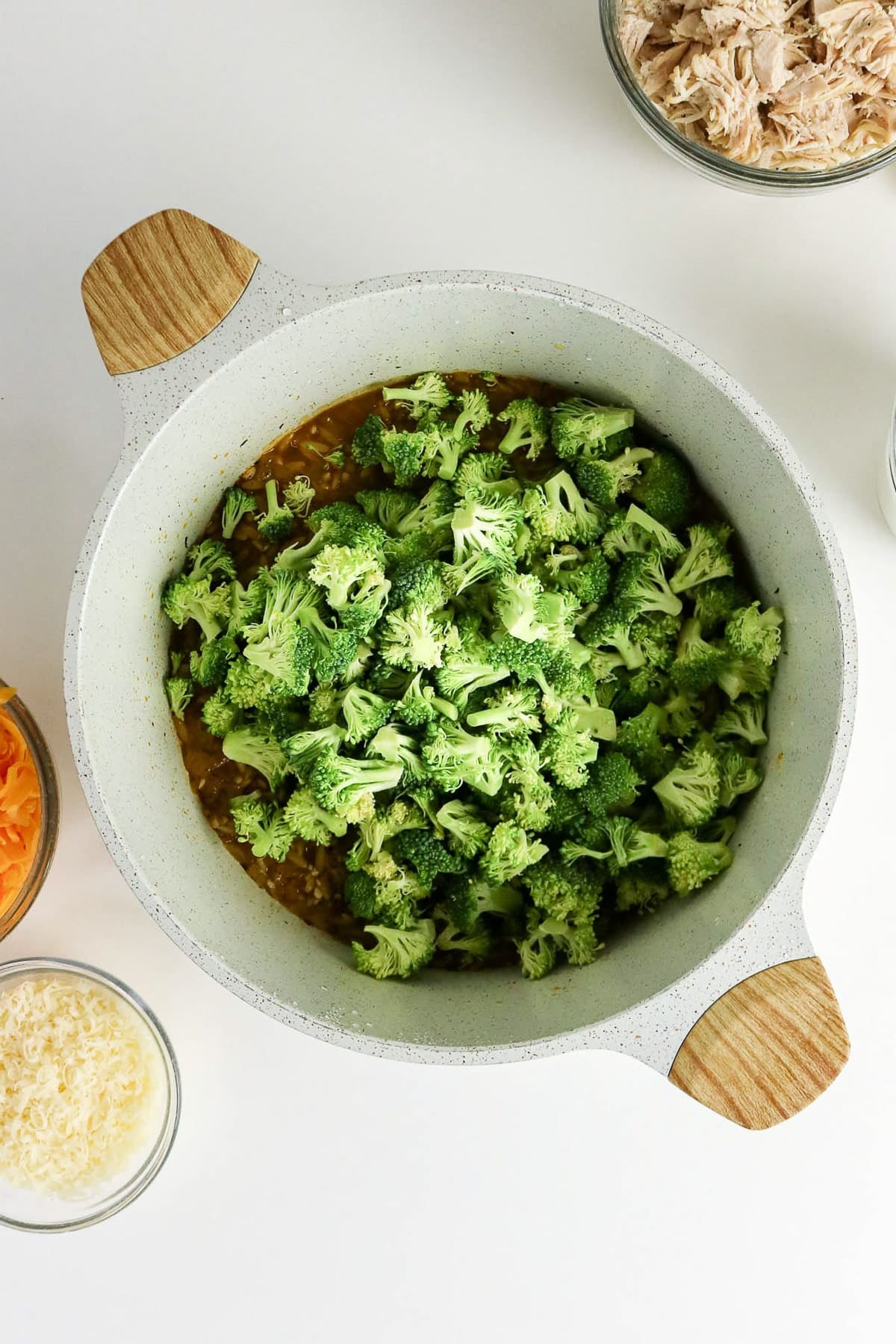 Overhead view of a pot filled with chopped broccoli, surrounded by bowls of cheese and shredded chicken.