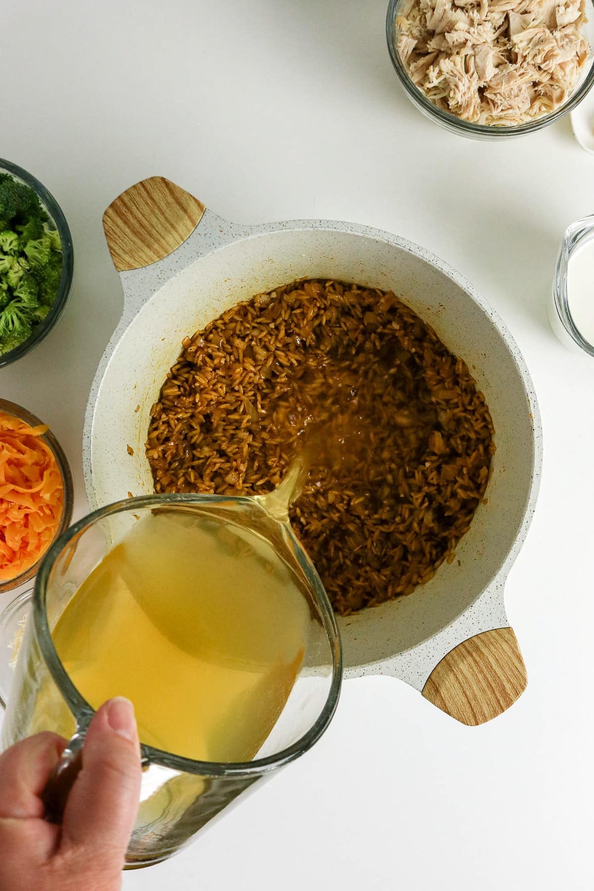 A hand pours broth into a pot of cooked rice, with bowls of chicken, cheese, and broccoli nearby.