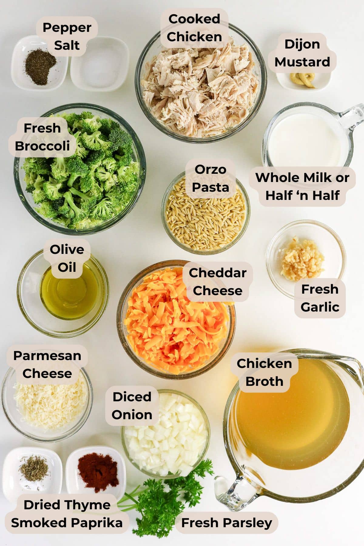 Bowls of shredded chicken, orzo, veggies, cheese, broth, milk, and spices arranged on a white surface.