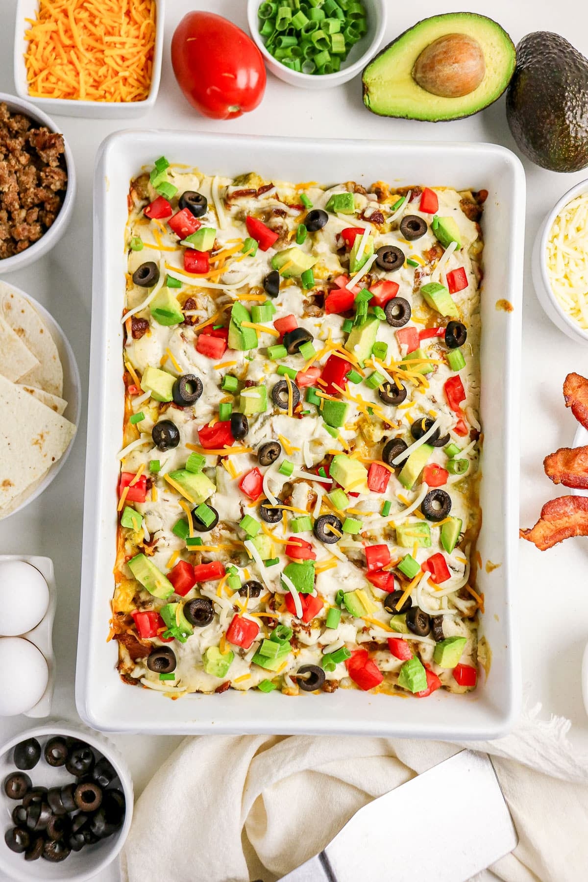 A casserole topped with cheese, olives, avocado, tomatoes, and green onions, surrounded by taco ingredients.