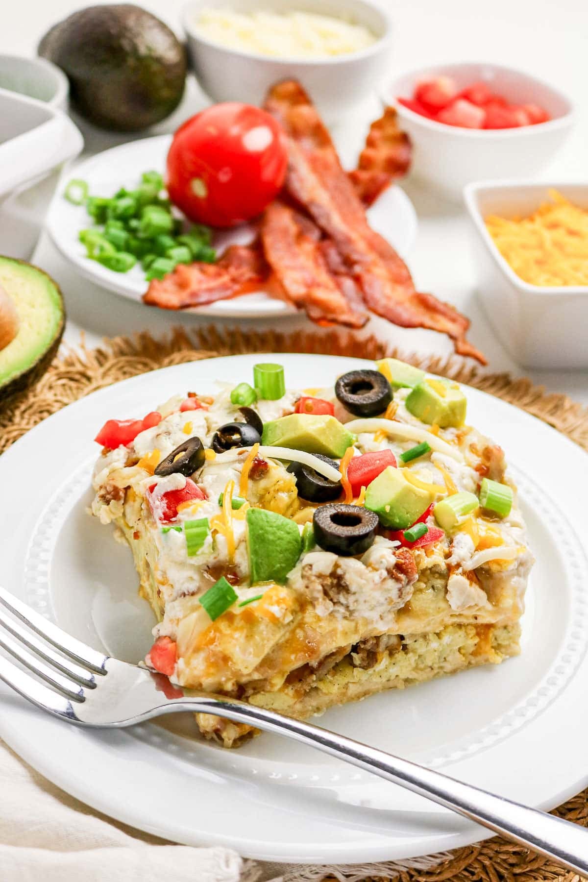A slice of breakfast casserole topped with veggies, served with bacon and fresh ingredients on the side.