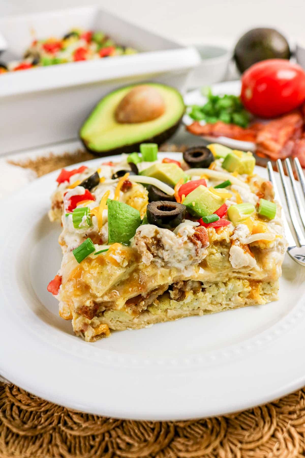 A slice of breakfast casserole topped with cheese, olives, tomatoes, and green onions on a white plate.