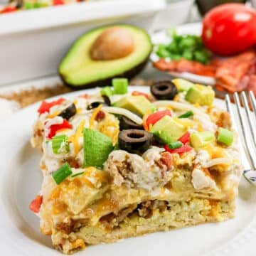 A slice of breakfast casserole topped with avocado, olives, cheese, green onions, and tomatoes on a white plate.
