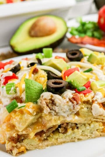 A slice of breakfast casserole topped with avocado, olives, cheese, green onions, and tomatoes on a white plate.
