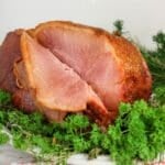 A sliced glazed ham on a plate, garnished with fresh green herbs.