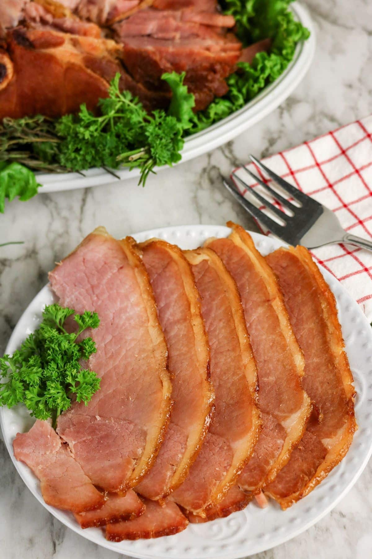Sliced ham on a white plate, garnished with parsley, with a serving fork and napkin nearby.