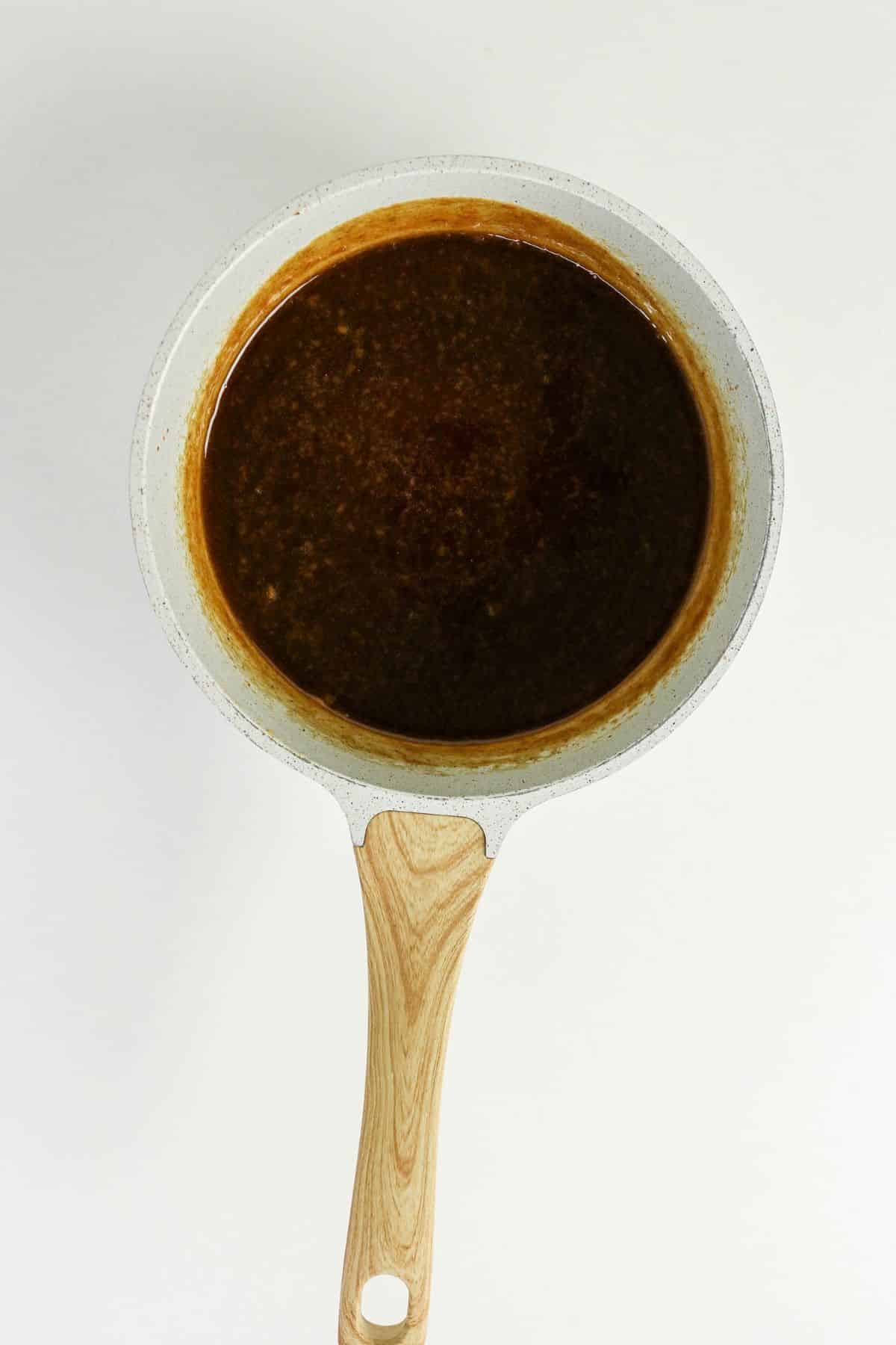 White saucepan with a wooden handle filled with brown sauce, viewed from above on a white background.