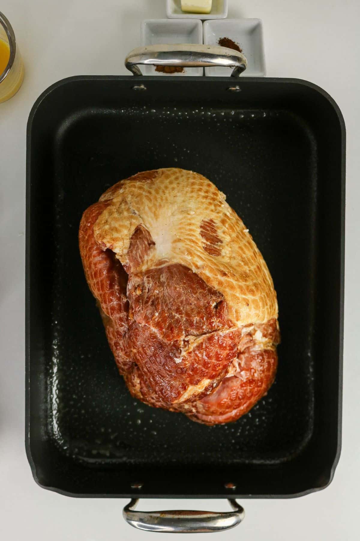 A raw ham sits in a black roasting pan on a white surface, ready to be cooked.