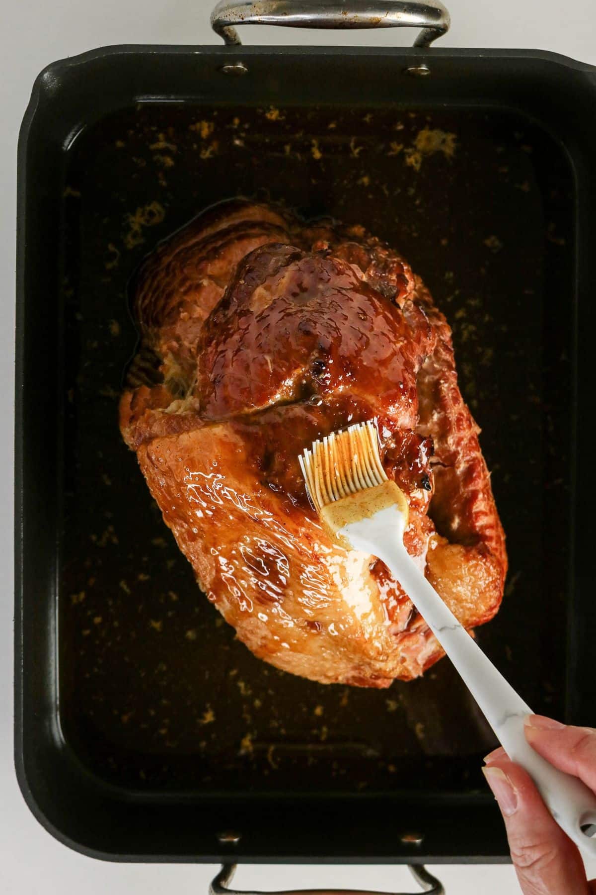 A hand brushes glaze onto a roast in a black roasting pan filled with juices.