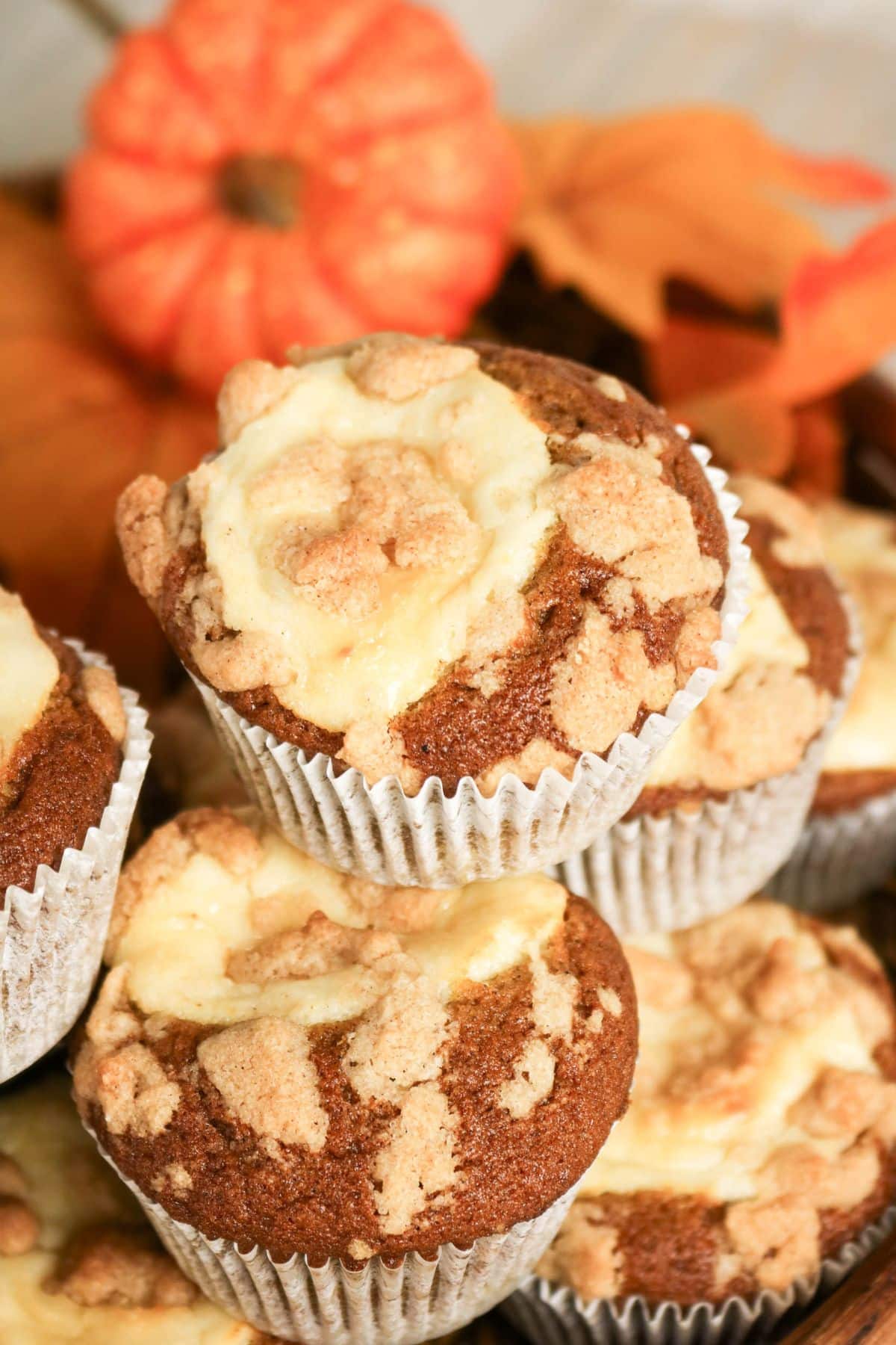Pumpkin muffins with crumb topping stacked, with a small pumpkin and autumn leaves in the background.
