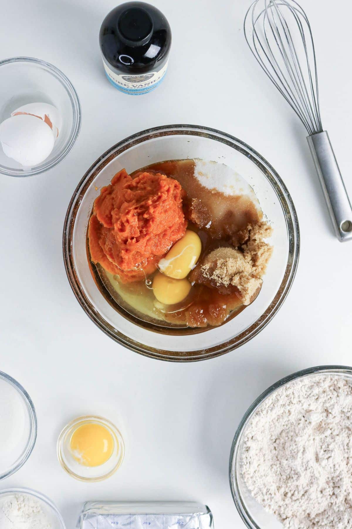 Glass bowl with pumpkin, eggs, sugar, and brown sugar, surrounded by flour, vanilla, and whisk on white surface.