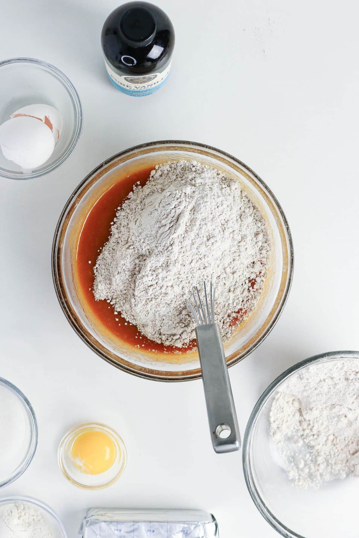 Mixing bowl with flour and wet ingredients, whisk, eggs, sugar, and vanilla bottle on a white surface.
