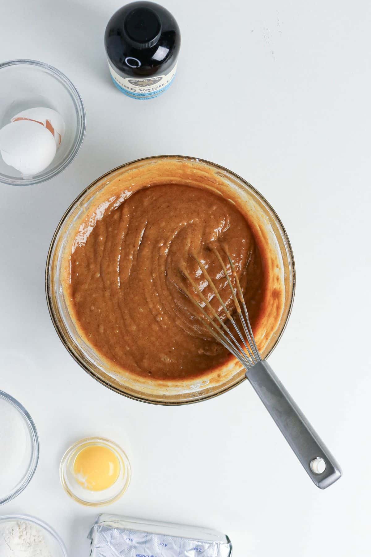 A bowl of brown batter with a whisk, surrounded by eggs, sugar, flour, and a bottle of vanilla.