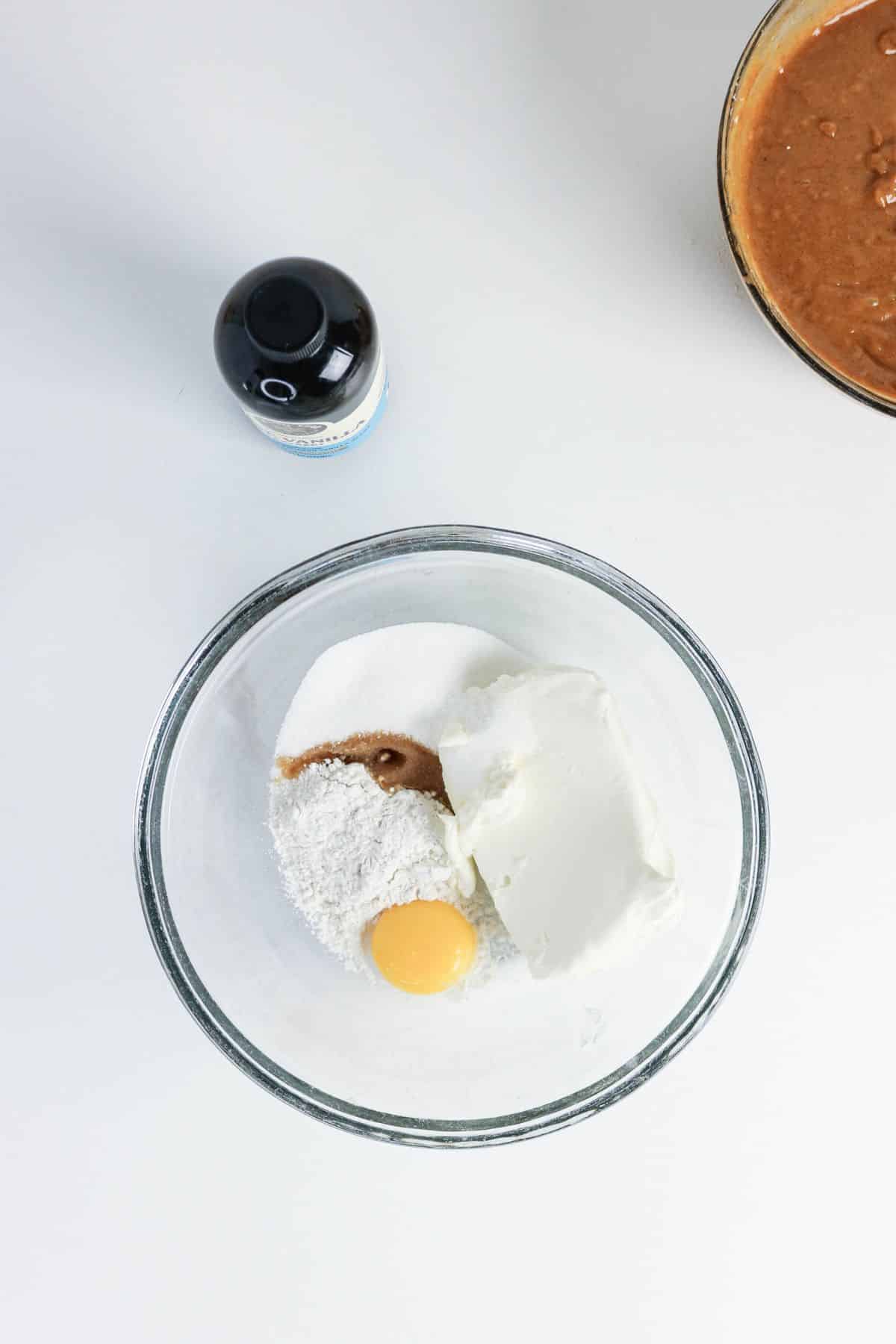 A glass bowl with cream cheese, egg yolk, flour, sugar, and vanilla; bottle and batter bowl nearby.