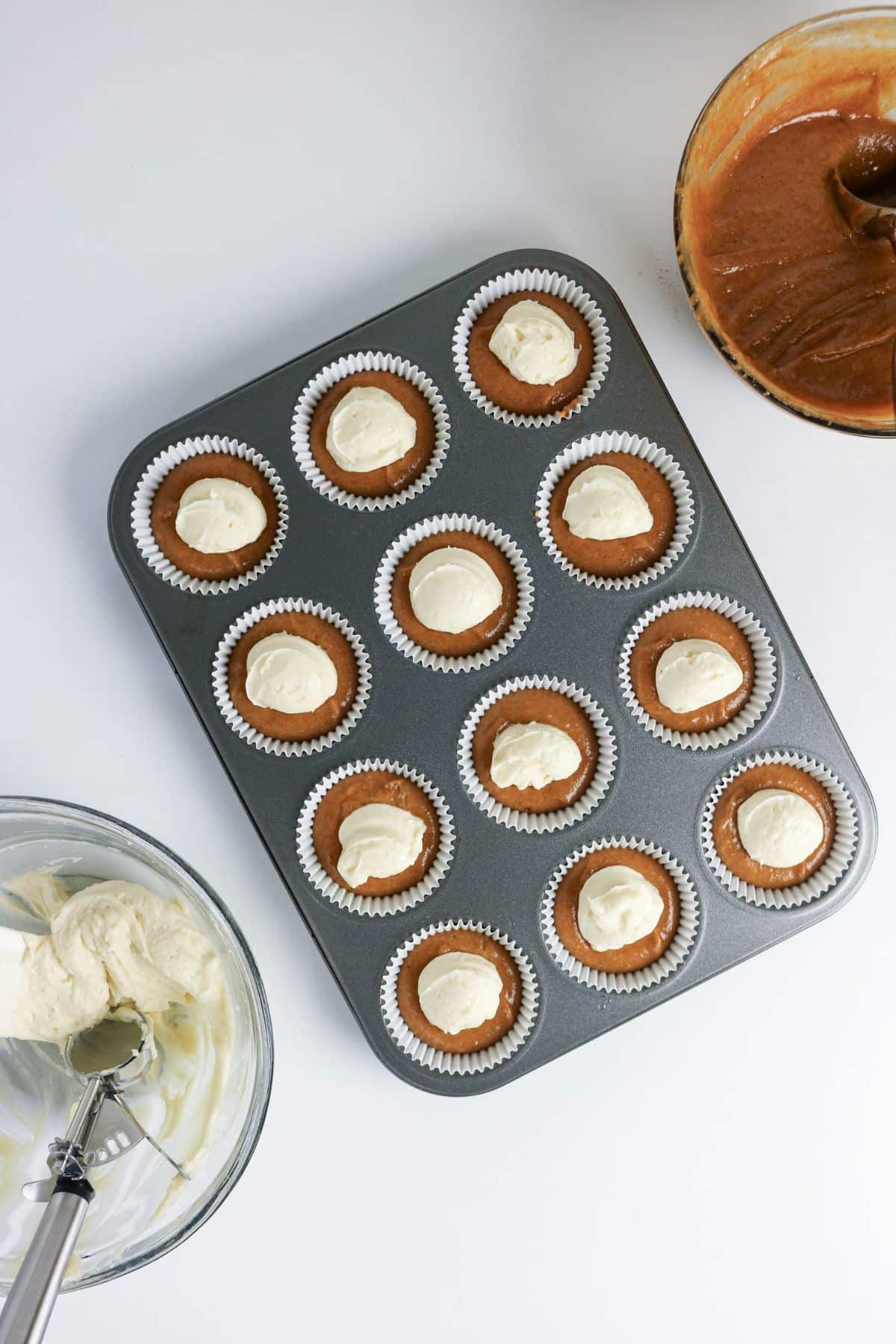 A muffin tin with batter-filled liners, each topped with a dollop of cream; bowls and scoop nearby.