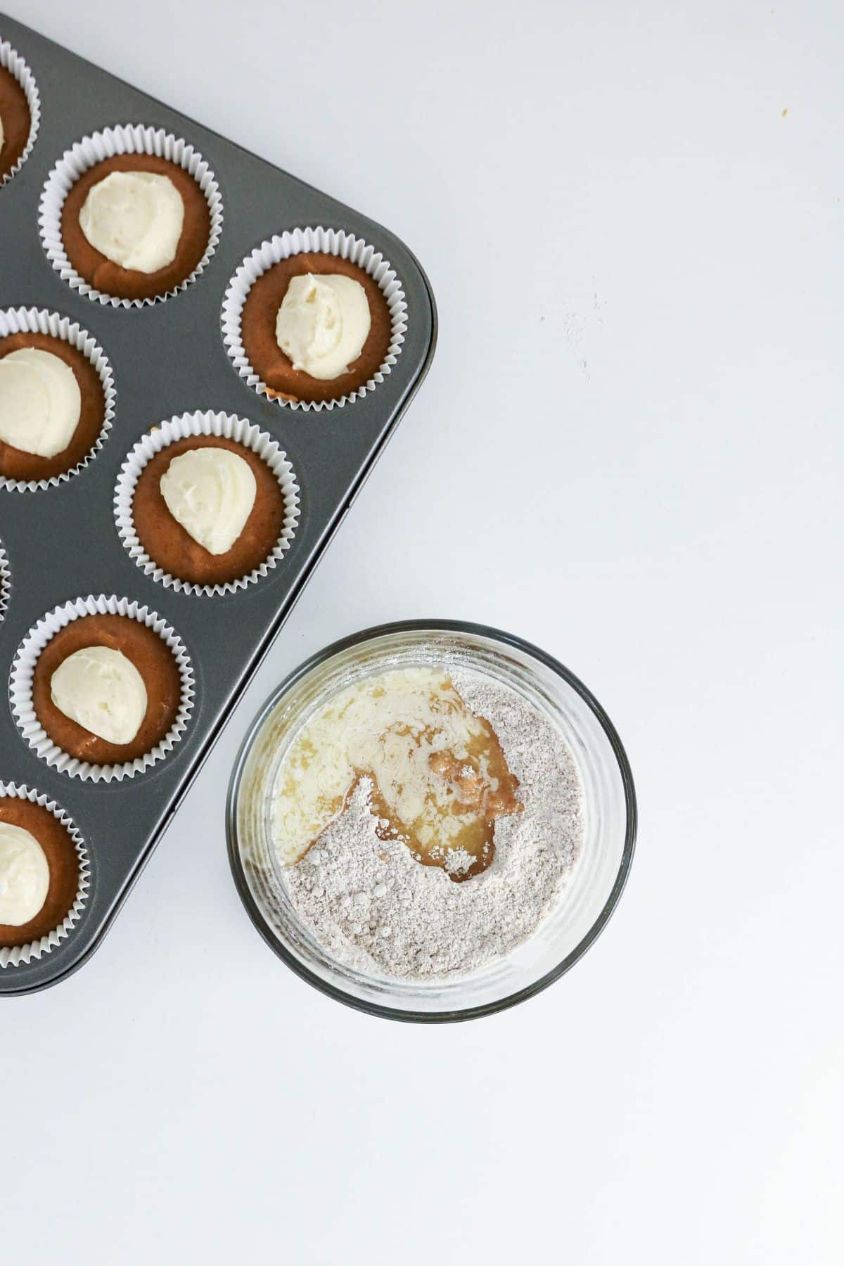 Muffin tray with batter beside a glass bowl of dry and wet baking ingredients on a white surface.