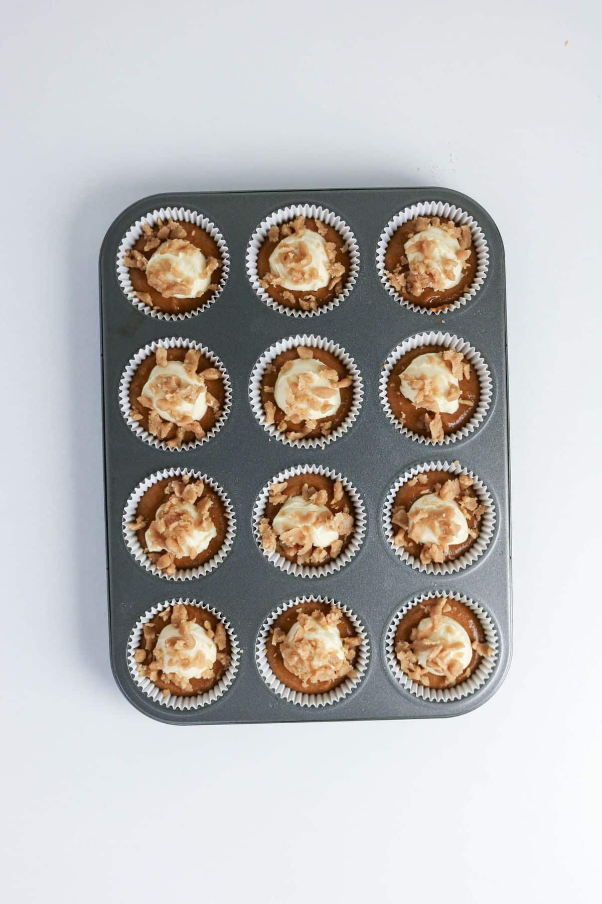 A muffin tin with twelve cupcakes topped with swirled frosting, viewed from above on a white background.