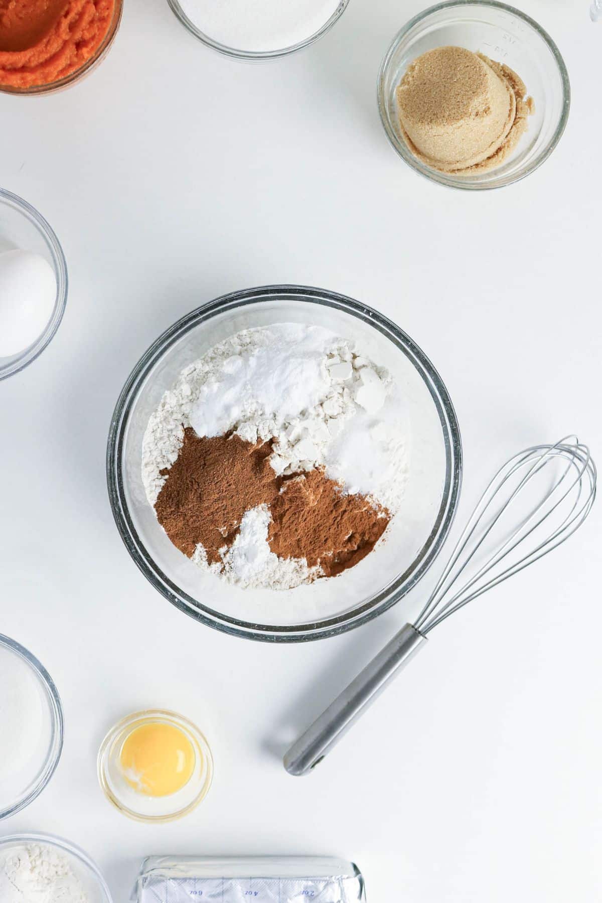A mixing bowl with flour, spices, and baking powder, surrounded by eggs, sugar, and a whisk on a white surface.
