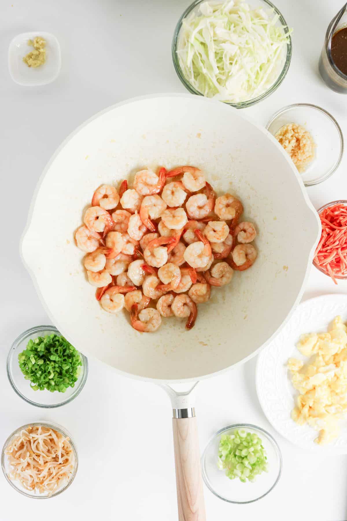 Cooked shrimp in a white pan surrounded by bowls of chopped cabbage, green onions, eggs, and seasonings.
