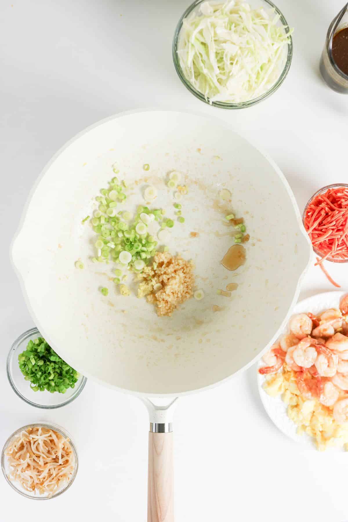 A white pan with garlic and green onions, surrounded by shrimp, shredded vegetables, and sauces on a white surface.