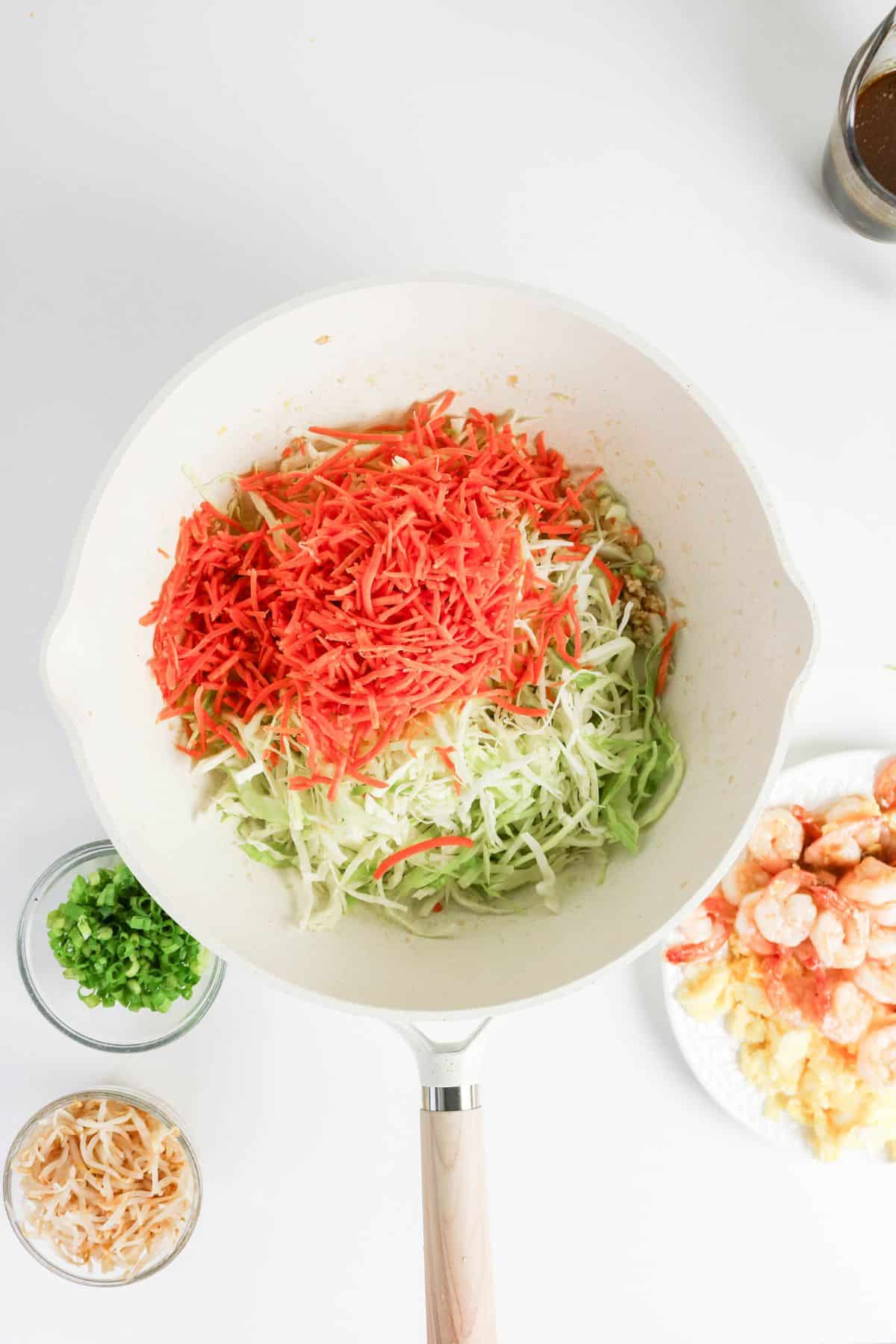 White pan with shredded carrots and cabbage, surrounded by small bowls of green onions, shrimp, and sprouts.