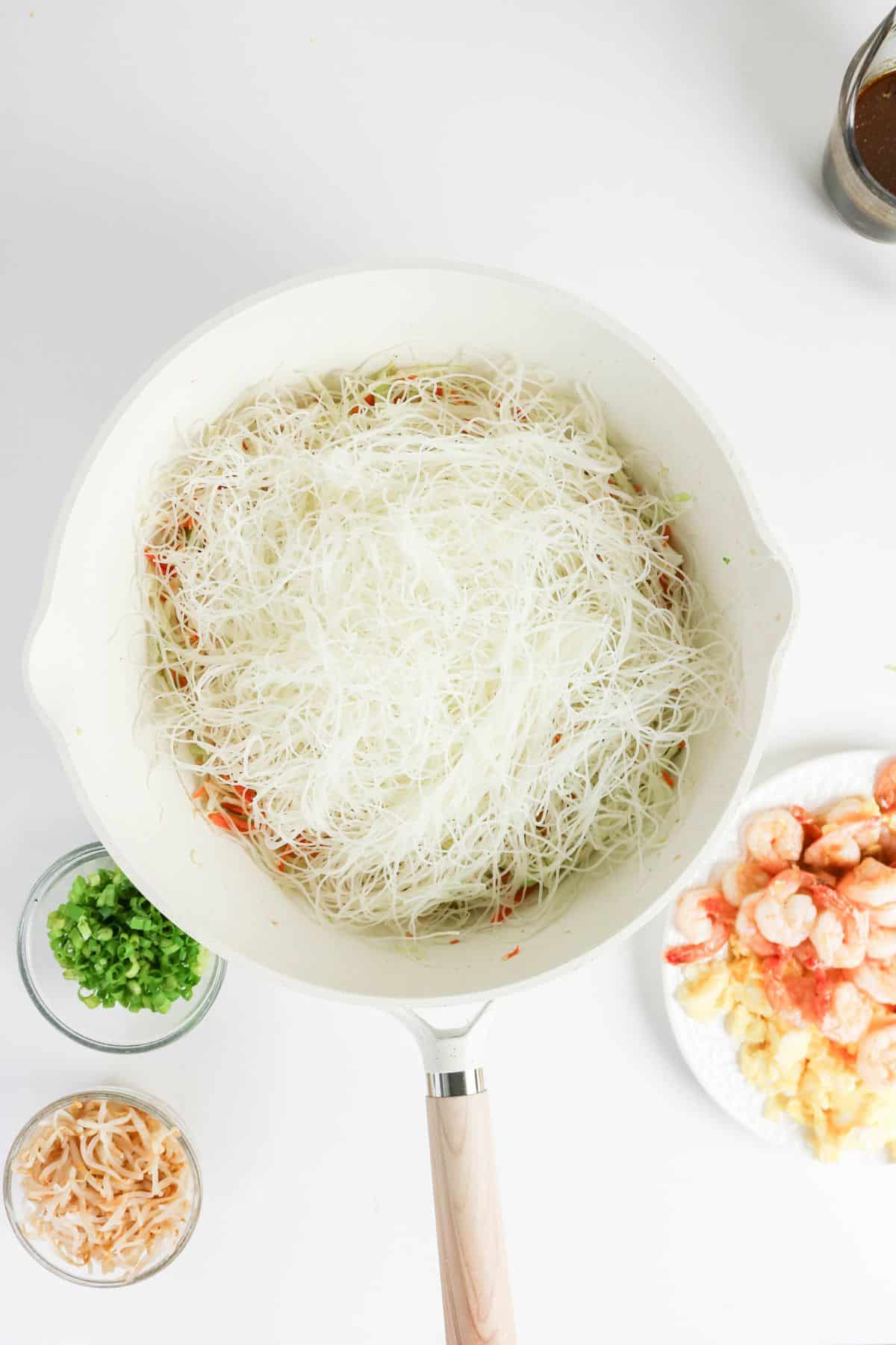 White rice noodles in a pan, with bowls of green onions, dried shrimp, and cooked shrimp beside it.