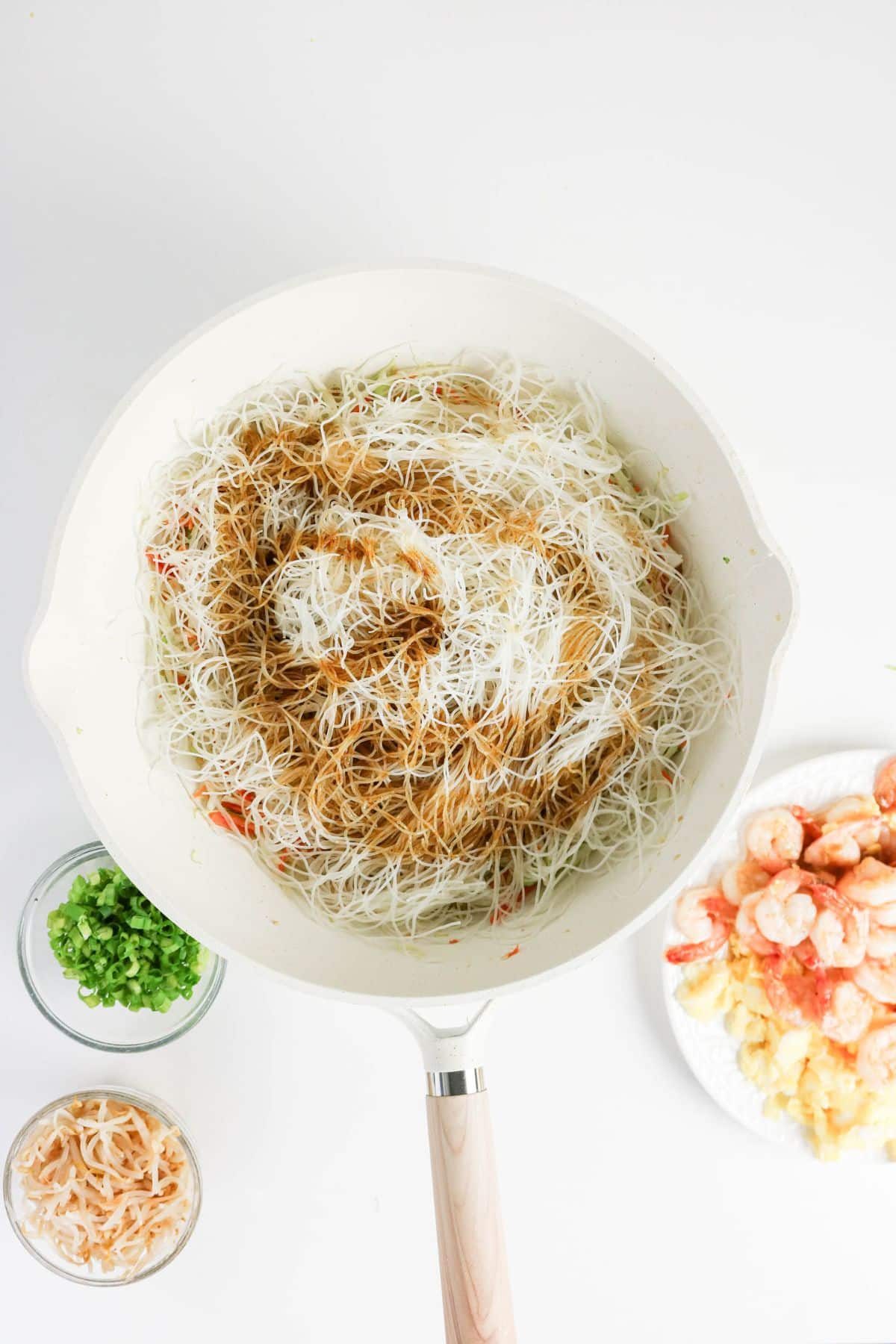 White pan with rice vermicelli, soy sauce, and vegetables, next to bowls of shrimp, green onions, and dried shrimp.