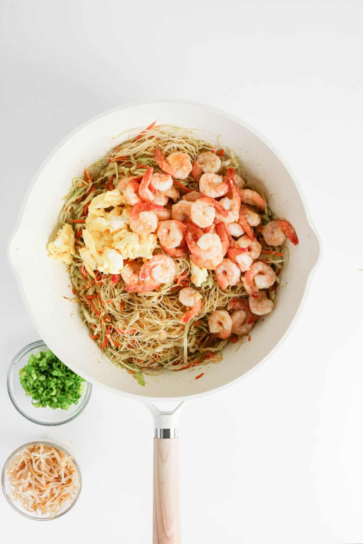 White pan with shrimp, scrambled eggs, and noodles, next to bowls of chopped green onions and shredded cheese.