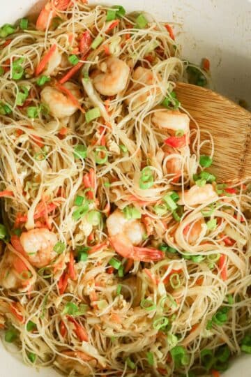 Rice noodles with shrimp, green onions, and shredded vegetables in a white bowl, mixed with wooden utensils.