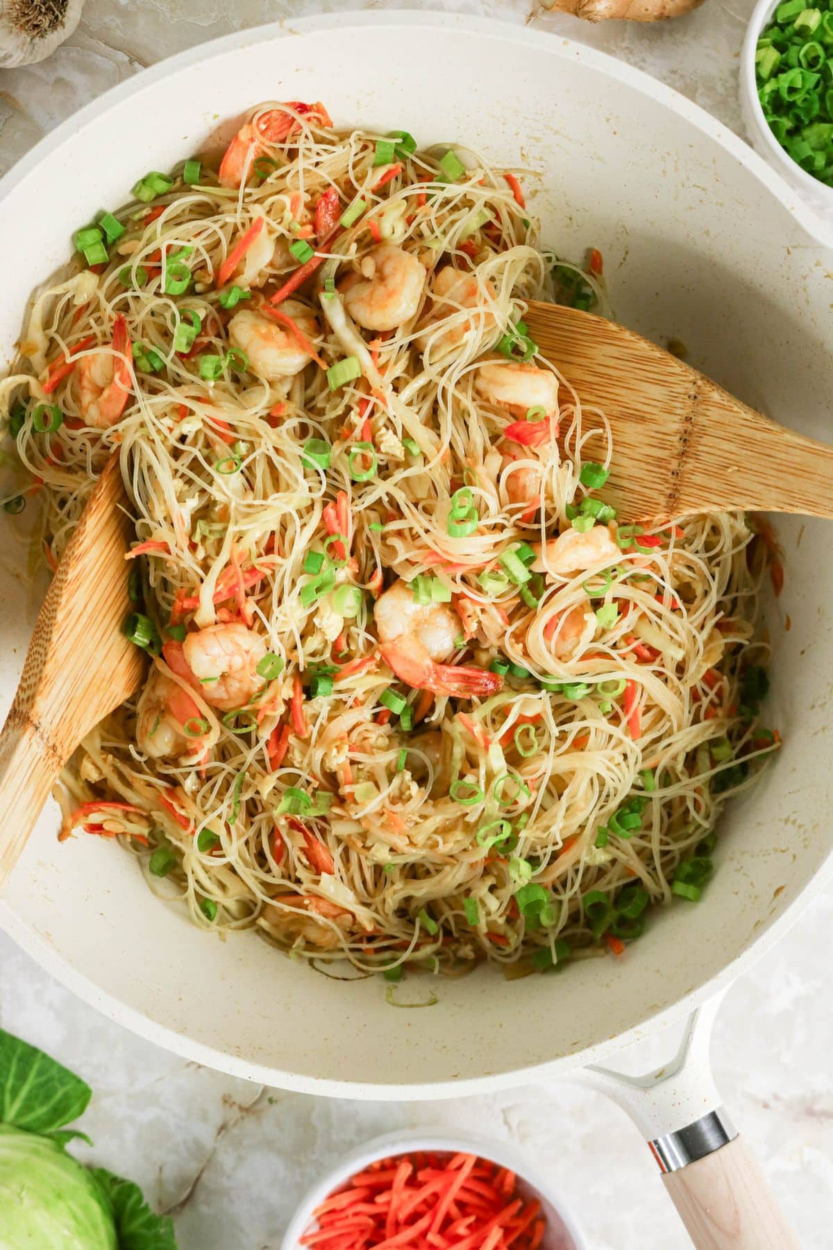 Rice noodles with shrimp, carrots, and green onions being mixed in a white pan with wooden utensils.