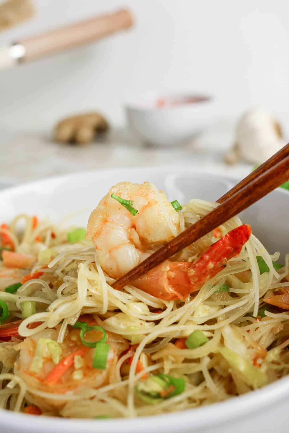 Chopsticks holding shrimp and noodles above a bowl of stir-fried noodles with vegetables.