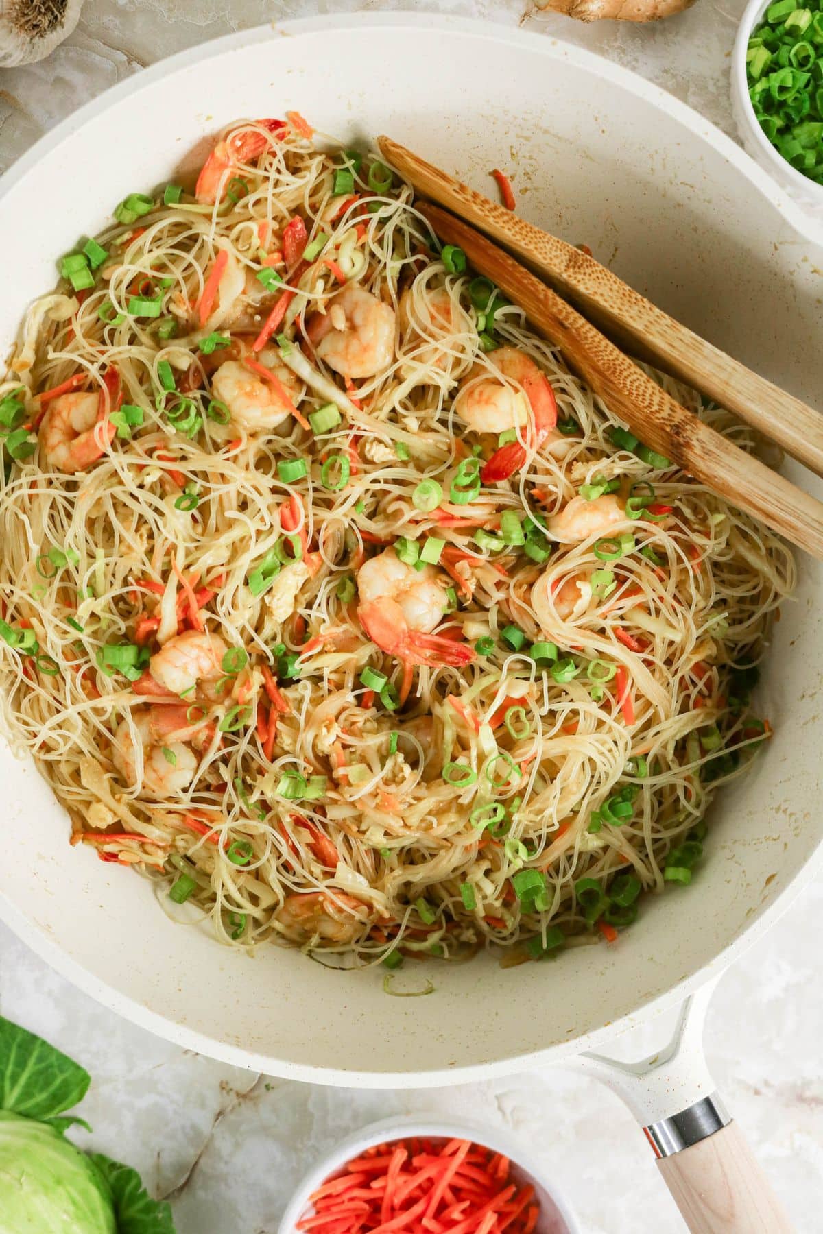 Rice noodles with shrimp, sliced carrots, and green onions in a white pan with wooden tongs.