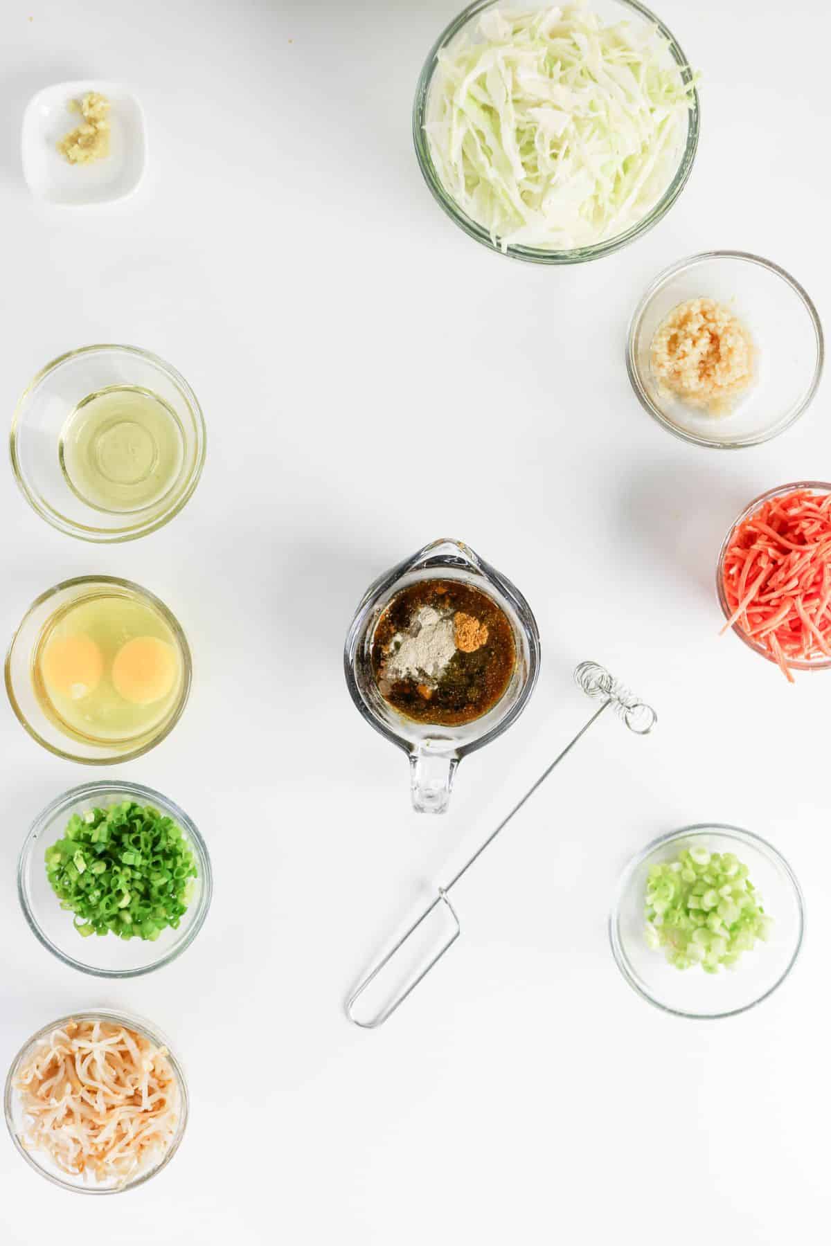 Various bowls with shredded cabbage, eggs, seasonings, and vegetables arranged around a whisk on a white surface.