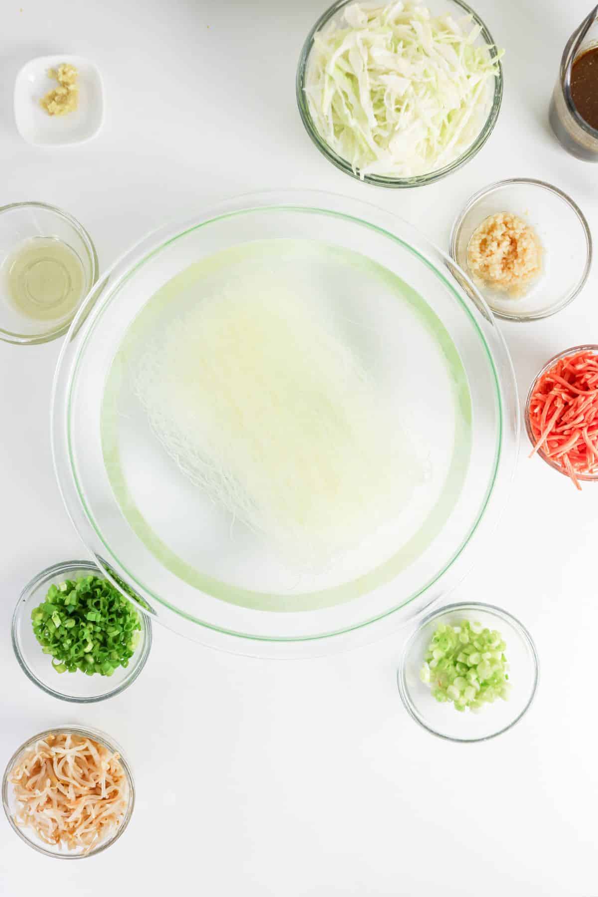 Glass bowl with uncooked vermicelli in water, surrounded by prep bowls of veggies and seasonings on a white surface.