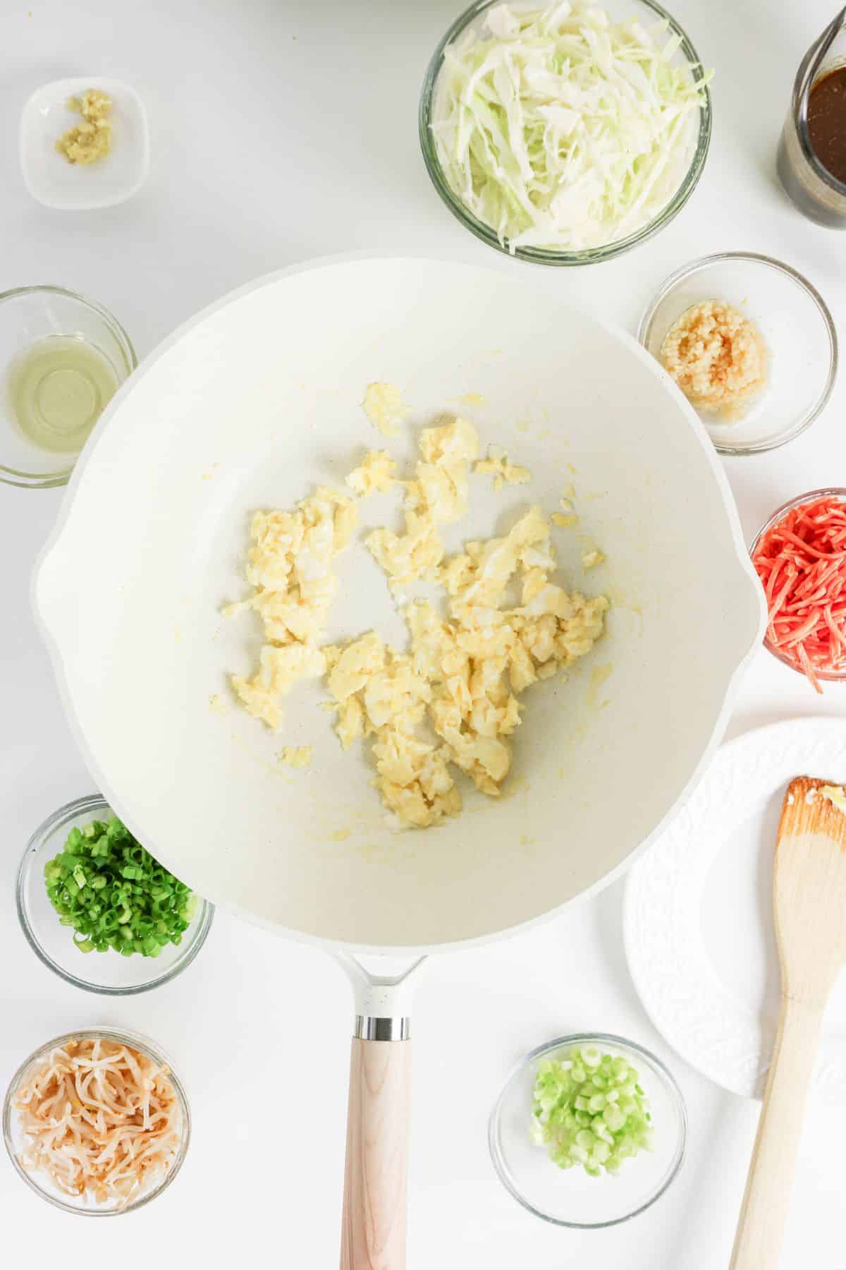 Scrambled eggs in a white pan surrounded by bowls of chopped vegetables and seasonings on a white surface.