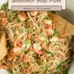 A bowl of Shrimp Mei Fun noodles with green onions and carrots, mixed gently with wooden utensils.