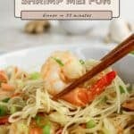 Chopsticks holding shrimp over a plate of Shrimp Mei Fun noodles with veggies and green onions.