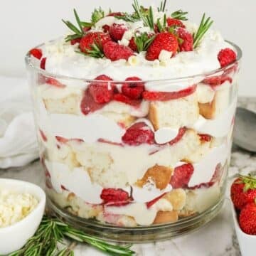 The finished trifle in a clear glass bowl, topped with strawberries, raspberries, white chocolate curls, and rosemary sprigs.