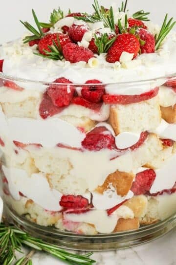The finished trifle in a clear glass bowl, topped with strawberries, raspberries, white chocolate curls, and rosemary sprigs.