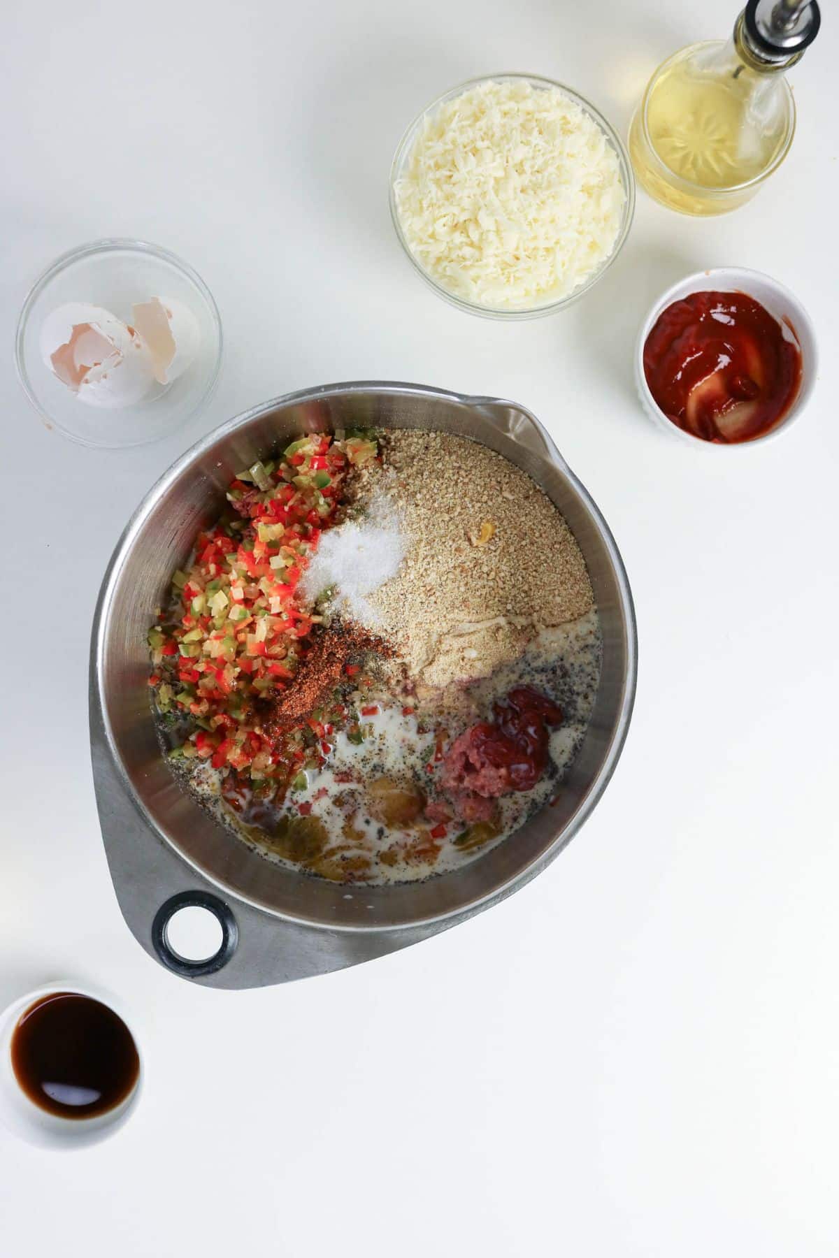 A metal mixing bowl containing ground meat, chopped vegetables, breadcrumbs, spices, and liquid ingredients.