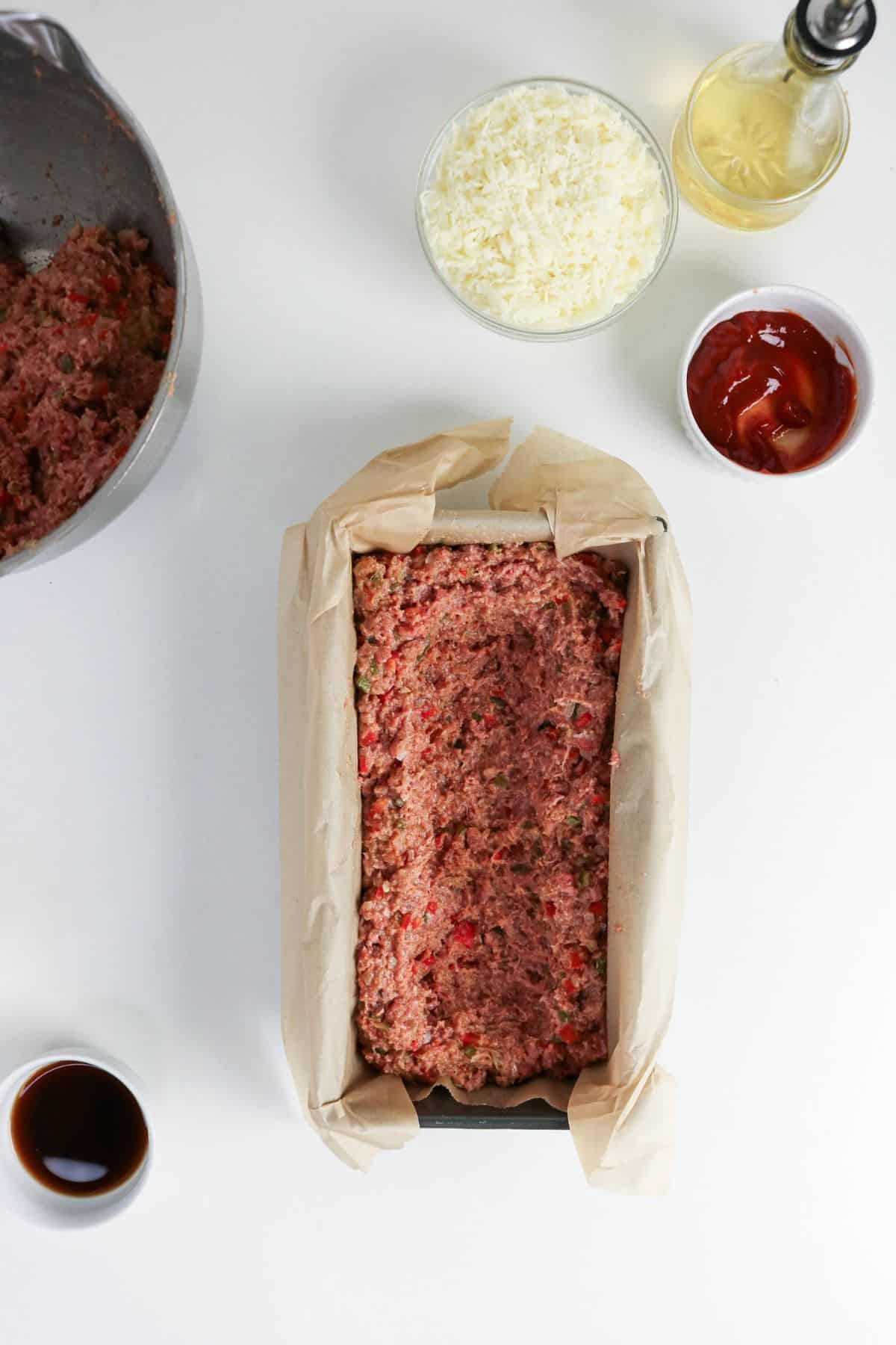 Raw meatloaf mixture in a parchment-lined loaf pan, with bowls of cheese, ketchup, and sauce nearby.